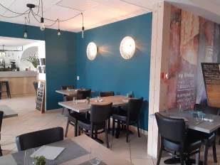 Photo de l'intérieure du restaurant nous y voyons de beaux lustres éclairant la pièce d'une lumière chaleureuse et de jolies tables dressées devant un mur peint d'une couleur bleu.