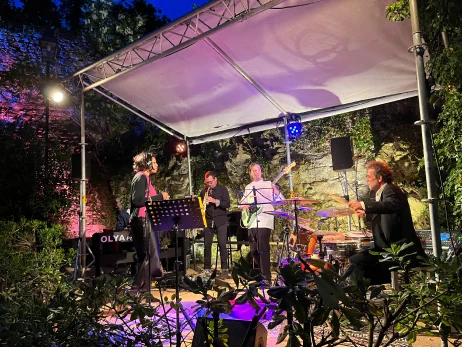 Scène de concert entouré de plantes dans la cour intérieure, avec sous une tonnelle des musiciens qui jouent et une chanteuse qui les accompagne.