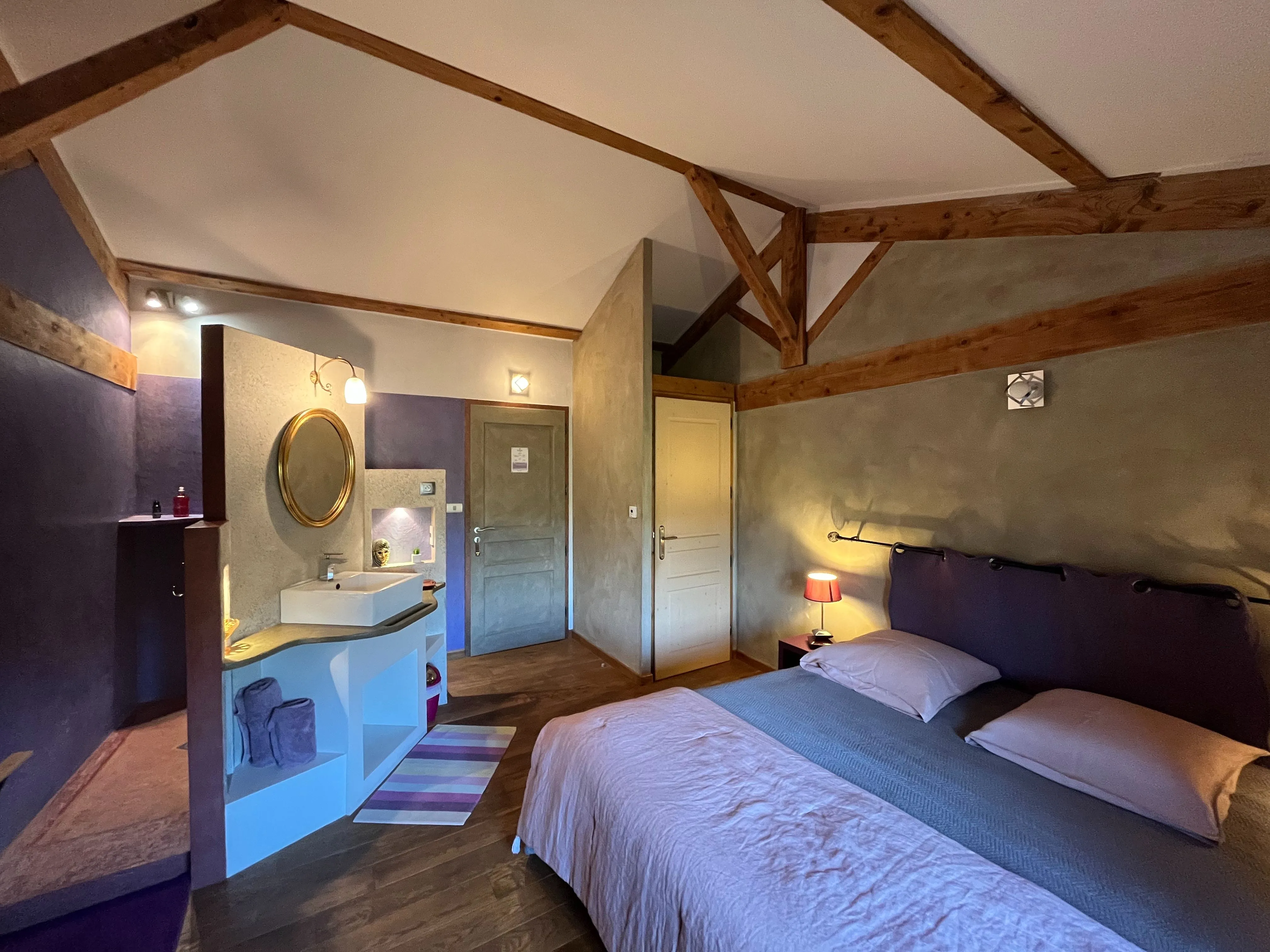 Toscaanse kamer - Overzicht van de kamer met tweepersoonsbed en aangeschakelde nachtkastlamp, centrale ingang, toiletdeur en grote tadelakt-douche met enkele wastafel