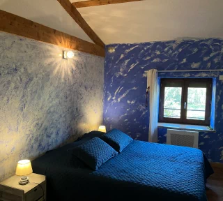 Chambre créole aux couleurs bleu, blanche et verte. Nous voyons un lit double. Ainsi qu'une fenêtre avec vue sur le jardin