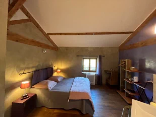 Chambre à thématique toscane, aux couleurs grise et violette. On aperçoit un grand lit double, une fenêtre avec vue sur le jardin ainsi qu'une table et des rangements.