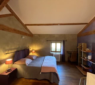 Chambre à thématique toscane, aux couleurs grise et violette. On aperçoit un grand lit double, une fenêtre avec vue sur le jardin ainsi qu'une table et des rangements.