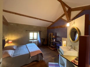 Chambre à thématique toscane, aux couleurs grise et violette. On aperçoit un grand lit double, une fenêtre avec vue sur le jardin ainsi qu'une table, des rangements et un lavabo.