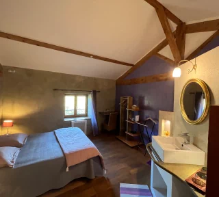 Chambre à thématique toscane, aux couleurs grise et violette. On aperçoit un grand lit double, une fenêtre avec vue sur le jardin ainsi qu'une table, des rangements et un lavabo.