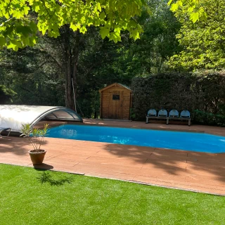 Une belle piscine bleue ombragée, entourée d'une terrasse en bois. Nous voyons quelques transats ainsi qu'un petit cabanon en bois en fond.